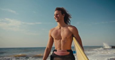 Kıvırcık saçlı, deniz kıyısında yürüyen ve yazın ellerinde sarı sörfünü tutan erkek sörfçünün ön görüntüsü..