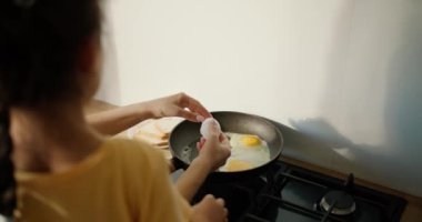 Sarı elbiseli esmer bir kızın, annesiyle birlikte modern bir mutfağın fırınındaki tavada kızarmış yumurta hazırlarken görüntüsü. Annesiyle birlikte kahvaltı yapmayı öğrenen bir kız.