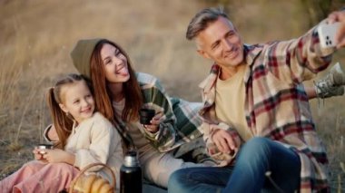 Orta yaşlı mutlu bir adam, karısı, yeşil kareli gömlekli esmer bir kız ve küçük bir kız, şehir dışında piknik yaparken beyaz bir telefonla selfie çekiyorlar.