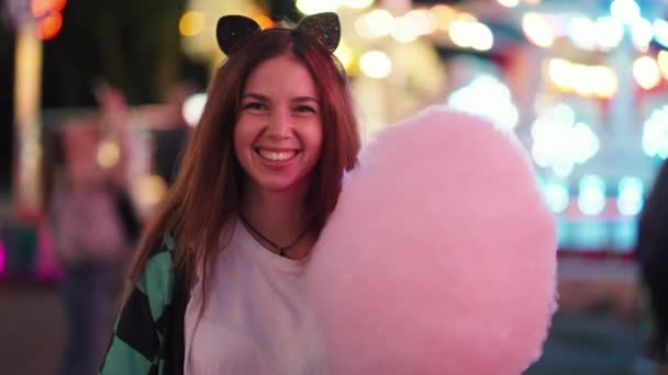 一个穿着一件白色T恤衫的黑发女孩在明亮的游乐园里咬着一个大的粉色棉花糖的画像 — 图库视频影像