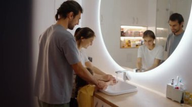 Sırtından vurulmuş, gri tişörtlü esmer bir adam, beyaz tişörtlü esmer bir kız, ellerini sarı elbiseli, modern lavabolu küçük kızlarıyla yıkıyorlar.