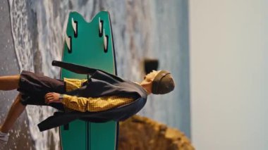 Kareli turuncu tişörtlü bir sörfçünün elinde sörf tahtasıyla kumlu bir sahil boyunca yürüdüğü dikey bir video..