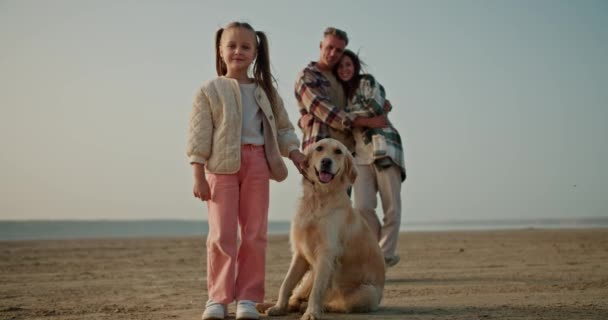 一个穿着白色夹克和粉色裤子的快乐的金发小女孩的画像 她站在她的大奶油色的狗旁边 身后是她快乐的父母 一个中年男人和一个穿着绿色衣服的黑发女孩 — 图库视频影像