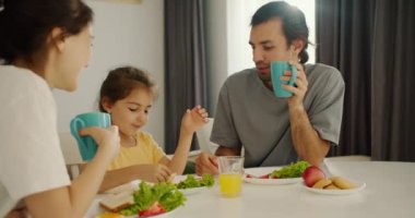 Gri tişörtlü esmer bir adam elinde turkuaz bir bardak tutuyor ve karısı ve küçük kızıyla kahvaltı ediyor. Modern mutfağın beyaz masalı sarı bir elbisesi var..