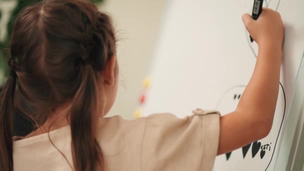 黒いマーカーを使った少女は 部屋のデッサンマグにイーゼルの上に立っている大きな白い紙を描きます 幼稚園の少女は大きなマーカーで描画する — ストック動画