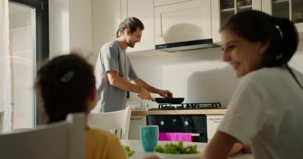若いブルネットの男は キッチンで朝食を準備しながら妻と娘のためにフライパンにトーストを投げることができる方法を示しています 男の妻と娘は彼が朝の準備をするのを見ている — ストック動画