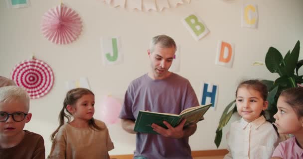 一个穿着紫色T恤 白发苍苍的男人和一个穿着白色衬衫 发型蓬松的女孩 与孩子们围坐在地板上看书 那个人在读一本学前班的书 — 图库视频影像