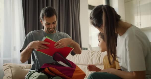 快乐的年轻父母们正在为女儿上学做准备 并为她打包背包 他们把不同的笔记本和课本放在一间现代房间的浅褐色沙发上 — 图库视频影像