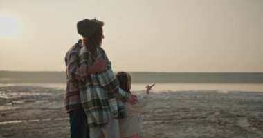 Kareli gömlekli, mutlu esmer bir adamın arka görüntüsü. Yaz akşamında şehir dışında, ıssız bir sahilde, karısına ve kızına sarılıyor..