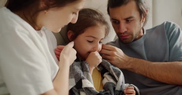二人の両親 灰色のTシャツのブルネット男と彼の妻 白いTシャツのブルネットの少女は ソファーの毛布に座っている病気の娘を世話し ストロークしています — ストック動画