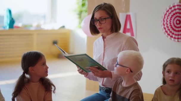 一位戴眼镜 身穿白色和粉色衬衫的金发女子为俱乐部的学生们读一本绿皮书 以帮助孩子们为上学做准备 — 图库视频影像