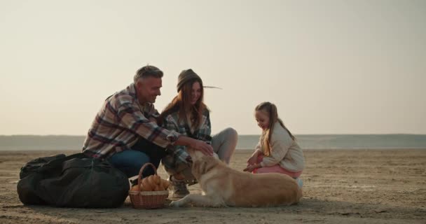 快乐的黑发男人穿着灰色的衬衫爱抚他的狗 夏天他和妻子 女儿在荒芜的海滨徒步旅行时放松下来 — 图库视频影像