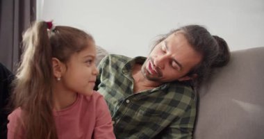 Kareli yeşil gömlekli esmer bir adam kanepeye oturur ve modern bir evde pembe ceketli esmer kızıyla iletişim kurar. Mutlu esmer adam, bekar baba.