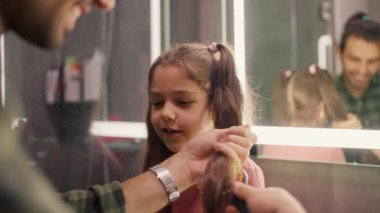 Yeşil kareli gömlekli mutlu esmer adam küçük kızlarının saçlarını tarıyor modern banyoda pembe bir elbise giymiş at kuyruklu esmer kız saç stili..