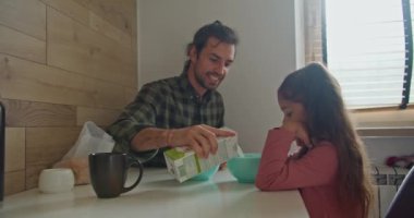 Yeşil kareli gömlekli mutlu esmer adam babası kendisi ve kızı için mavi tabaklara süt döker. Mutfaktaki masada kahvaltı yaparken pembe elbiseli esmer kız..