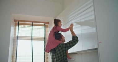 Pembe elbiseli esmer bir kız ekose gömlekli esmer babasına mutfaktaki üst raflardan bir şey almasında yardım ediyor. Küçük bir kız babasının omuzlarına oturur ve bir objeyi alır.