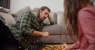 Yeşil kareli gömlekli esmer adam. Yalnız bir baba pembe ceketli esmer kızıyla satranç oynar. Modern bir dairede gri bir kanepeye yaslanır..