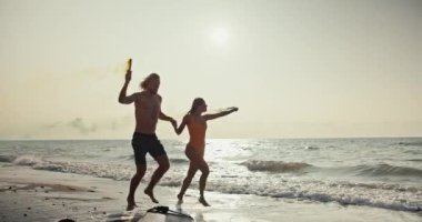 Sarışın bir adam turuncu mayo giymiş sarışın kız arkadaşını tutuyor ve ellerinde yeşil ve sarı renkler bırakan dumanlı havai fişekleri tutarak turuncu-kahverengi deniz kıyısında koşuyorlar.