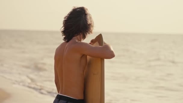 裸の胴を持つ長い髪のブルネット男の後ろの景色は 彼の手でサーフボードを保持し 朝の海岸で風に揺れている彼の髪をまっすぐにする — ストック動画