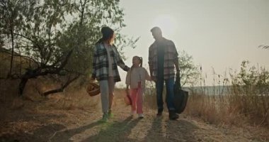 Yeşil kareli gömlekli esmer bir kız, sepetle birlikte, küçük kızı ve kocasıyla el ele yürüyor, gri saçlı, kareli gömlekli bir adam, piknikte
