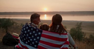 Amerika Birleşik Devletleri bayrağına sarılı mutlu bir aile yazın şehrin dışında piknik yaparken bir hasırın üzerinde oturup güzel bir akşam gün batımını izler..