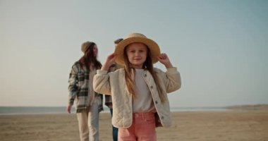 Hasır şapkalı mutlu küçük bir kızın portresi kameraya bakıyor ve gülümsüyor ve ailesi arkasından ona sarılıyor, ıssız bir sahilde piknik yaparken onunla poz veriyor.