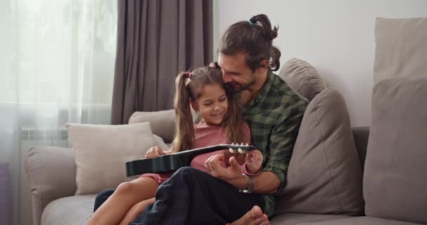 一个穿着粉色衣服的黑发小女孩坐在她的黑发父亲的腿上 穿着绿色格子衬衫 弹奏着四弦琴 坐在家里灰色的沙发上 — 图库视频影像
