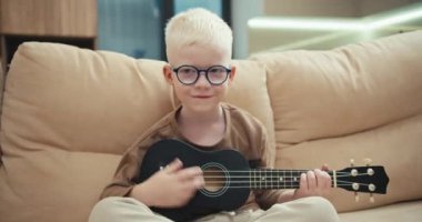 Beyaz saçlı, mavi yuvarlak gözlüklü, siyah ukulele çalan ve modern bir dairede krem rengi bir koltukta oturan mutlu bir albino çocuğun portresi..