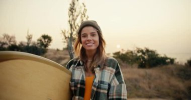 Ellerinde ahşap bir sörf tahtası tutan ekose gömlekli mutlu sarışın bir kızın portresi. Gülümsüyor ve kameraya bakıyor denizin, ağaçların ve çalıların arkasında.