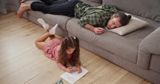一个穿着粉色连衣裙的金发小女孩躺在木地板上 画在一张白纸上 而她的单身父亲穿着绿色格子衬衫睡在灰色沙发上 — 图库视频影像