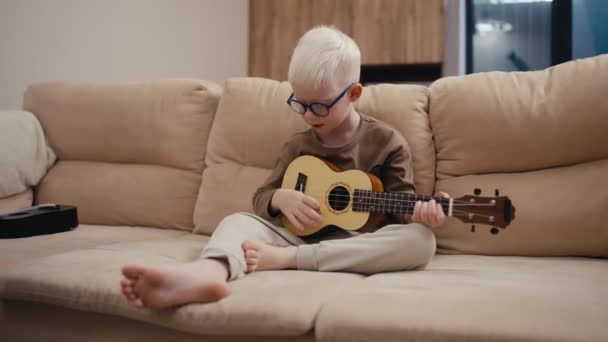 Мальчик Альбинос Белыми Волосами Круглых Синих Очках Играет Укулеле Сидя — стоковое видео