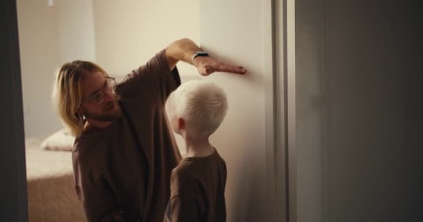 白い髪の小さなアルビノの少年は壁の近くに立って 彼のブロンドの父親はひげと眼鏡で彼の身長を測定し 次に彼に少年たちの身長を示しています あなたの子供の成長を測定する — ストック動画