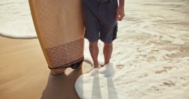 Yukarıdan yakın çekim, mavi şortlu bir adam sarı ahşap bir sörf tahtası tutuyor ve deniz kıyısındaki turuncu kumun üzerinde duruyor. Dalgalar ayaklarına geliyor ve sabah etraflarını sarıyorlar..