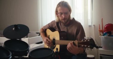 Kahverengi tişörtlü, sakallı sarışın bir adam yanında sarı akustik gitar çalıyor. Evde yetenek ve hobilerin davul seti gelişimi var..