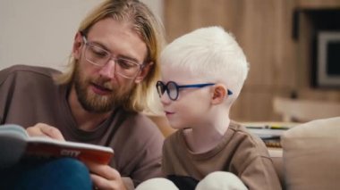 Babasıyla birlikte yuvarlak mavi gözlüklü, küçük bir albino çocuğun yakın çekimi, sakallı sarışın bir adam, ev ödevini yapıyor ve ona modern bir şekilde yazmayı ve okumayı öğretiyor.