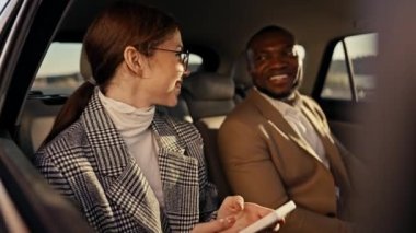 Yuvarlak gözlüklü mutlu bir esmer kız, modern bir arabada iş gezisi sırasında kahverengi takım elbiseli siyah bir adamla iletişim kurarken sevinir ve eğlenir..