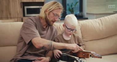 Gözlüklü, sakallı sarışın bir adam küçük oğluna mavi gözlüklü, beyaz saçlı albino bir çocuğun nasıl çalıştığını ve modern bir dairede müzik öğrenirken tellere işaret ettiğini anlatıyor..