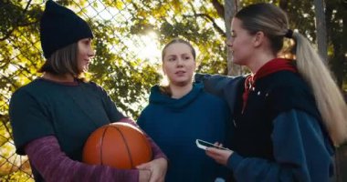 Spor giyimli mutlu üç sarışın kız iletişim kuruyor ve yaz aylarında yeşil ağaçların yanında turuncu bir basketbol topu tutuyor..