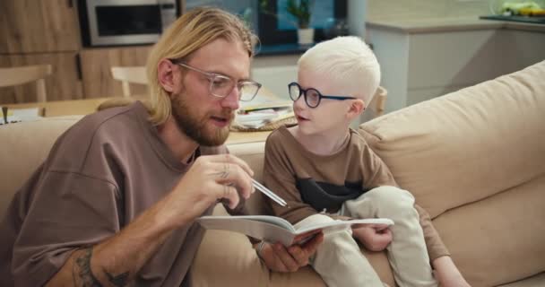 快乐的父亲 一个戴眼镜 留着胡子的金发男人 正在向他的小儿子解释如何在小学期间阅读和完成作业 他的小儿子是一个白化病男孩 戴着蓝色眼镜 头发是白的 — 图库视频影像