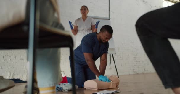 ボトムビュー 黒人の男性医師が医療用マネキンで人工呼吸を行い 彼の助手の少女看護師は白いレンガの壁に対して一般に説明します — ストック動画