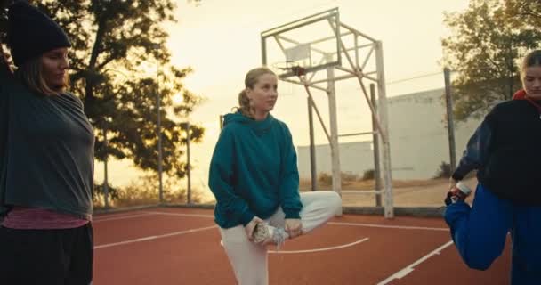 一个夏天的早晨 三个穿着运动服的女孩在红色运动场上打篮球前热身 她们在橙色篮球场附近的栅栏围起来打篮球 — 图库视频影像