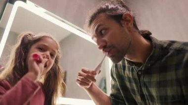 A little brunette girl with a ponytail and a pink dress brushes her teeth with a toothpick along with her single father, a brunette man there in a Green checkered shirt near a mirror in a modern