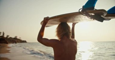 Rear view of a blond male surfer with a naked torso walking along the sandy seashore and carrying a surfboard above his head in the morning at sunrise. A professional surfer goes to the sea to ride