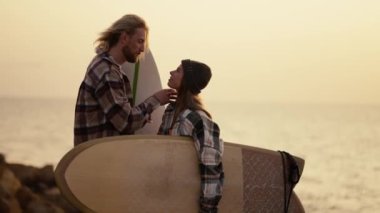 A blond guy with a beard in a plaid shirt straightens his hair and strokes the head of his blonde girlfriend in a black hat and plaid shirt who is holding a surfboard standing on a rocky shore near