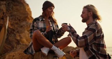 Happy couple blonde girl in a checkered shirt gives her boyfriend a mug of hot tea from a thermos and kisses him. A blond guy with a beard in a checkered shirt looks at his girlfriend, takes tea and