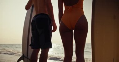 Zoom in: A brunette guy in shorts and a blonde girl in an orange swimsuit stand next to each other and hold surfboards and then they hold hands and look towards the sea.