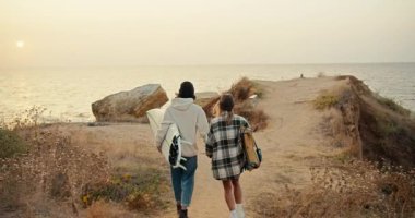 A happy brunette guy in a black hat in a white sweatshirt and his blonde girlfriend in a checkered shirt walk down the slope to the sea and hold surfboards in their hands near the sea in sunny weather