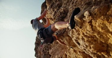 Shot from below, a man in a gray T-shirt and gray pants in a special insurance climbs a yellow steep rock and stones against the backdrop of a sunny clear summer sky.