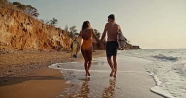 A happy couple, a blond guy and his blonde girlfriend in an orange swimsuit, walk and hold hands along the rocky sandy shore of the sea, they hold surfboards in their hands and walk along small waves