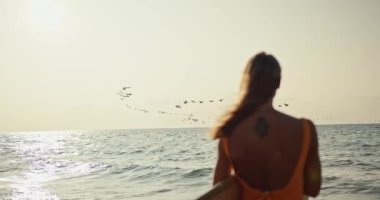 Rear view of a blonde girl in an orange swimsuit standing with a surfboard on the seashore and looking at the sea and the sky where a large flock of birds are flying at Sunrise in the morning.